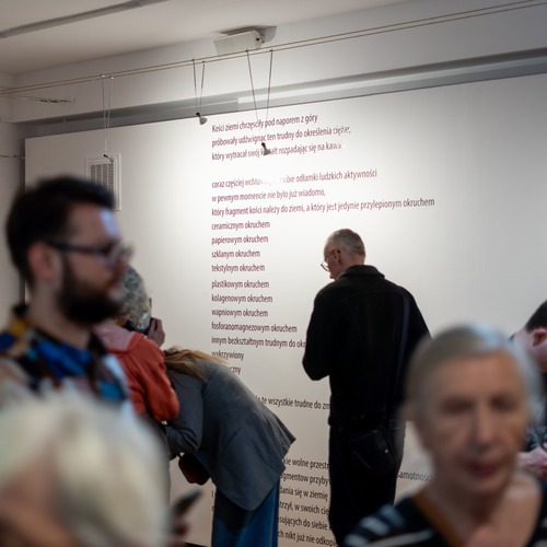 Zwiedzający w pomieszczeniu galerii, w tle biała ściana z czarnymi napisami.