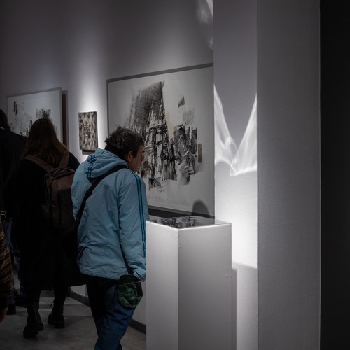 Zwiedzający w ciemniejszej sali galerii uważnie przyglądają się czarno-białym pracom i instalacjom artystycznym.